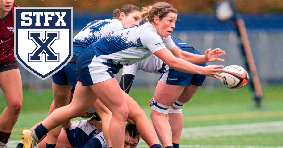 A StFX women's rugby player in white and navy passes the ball out of a ruck in the rain, with the StFX crest in the corner